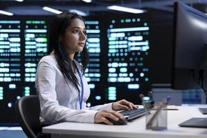 Indian woman doing yearly checkup to prolong data center electronics life span and avoid downtime. IT employee in server room monitoring server infrastructure components using PC photo