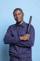 African american security guard feels proud with his police weapon on camera, posing with confidence with his baton and professional uniform. Trained bodyguard against blue background. photo