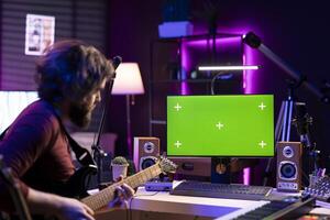 Young musician learning to play acoustic guitar with online lessons via computer with greenscreen layout in home studio. Audio engineer playing instrument to learn new accords, music industry. photo