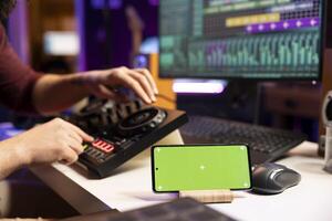 Artist engineer does mixing and mastering techniques on tunes, twisting pre amp knobs in home studio. Musician editing songs next to a smartphone with greenscreen display, daw software. photo