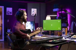 Beginner musician learning to tune his strings accords by watching tutorials online, twisting the knobs. Songwriter playing acoustic guitar in home studio, practicing musical instrument. photo