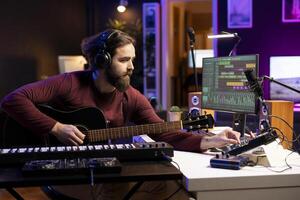 Singer creating tunes on guitar and adding sound effects in post production, uses mixing console and gear. Musician producing and editing audio recordings, playing acoustic instrument. photo
