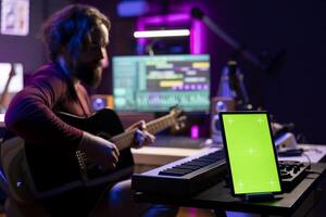 Singer producer playing acoustic guitar looking at daw software interface to record new tunes, learning accords on instrument strings. Composer singing songs next to greenscreen on tablet. photo