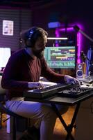 Songwriter plays midi controller piano notes in his home studio, using daw software on computer to record audio files. Young musician playing tunes on electronic keyboard, post production. photo