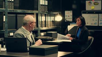 Senior detective asking his partner about legal documents in archive room, looking into crime trends and statistical data to connect different cases. Inspector examines legal statements. Camera A. photo