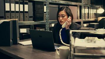 Law officer verifying details for the private investigation, working in archive room. Detective cross checks information on landline phone call, contacting people from records. Camera B. photo