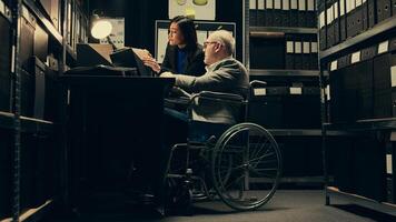Inspector consulting with criminal expert on archived materials and autopsy records, examining folders and old cases to find connections. Private investigation in disability friendly space. Camera B. photo