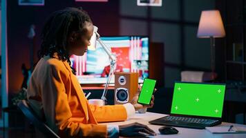 African american girl uses smartphone and pc with greenscreen, looking at modern devices running the same isolated mockup template on display. Young woman checks copyspace screens. Camera A. photo