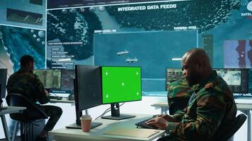 Military specialist monitors satellite data next to green screen display, working at command post with high tech gear to support army defense operations. Coordinates strategic missions. Camera B. photo
