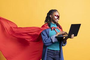 Happy girl portraying superhero with cape hiding identity, working on laptop, studio background. Woman wearing cloak posing as hero in costume, typing on notebook keyboard, covering face with mask photo