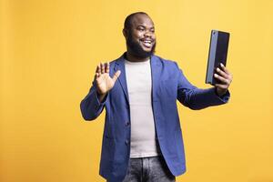 Jolly man excited to greet friends during teleconference meeting using tablet, studio background. Merry person waving hand, saluting mate during internet call on portable device photo