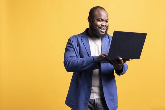 Cheerful man using laptop to do business related tasks for company, satisfied with profits, studio background. Joyful employee doing financial analysis on notebook, feeling pleased photo