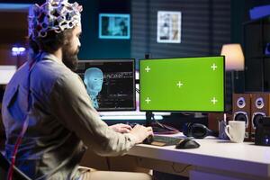 IT technician using EEG headset device translating thoughts into commands on green screen PC display on desk. System administrator controlling chroma key computer functions using mind photo