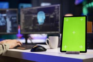 Focus on isolated screen tablet next to man in blurry background talking with AI gaining consciousness. Chroma key device on desk next to programmer, chatting with artificial intelligence, close up photo