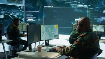 Military personnel in a base of operations using advanced computers and big screen, monitoring satellite systems to enhance security measures and process data. High tech army gear. Camera B. photo