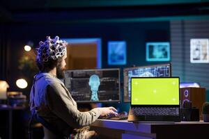 Software engineer with EEG headset starting mind upload trying to gain bionic powers, isolated screen on laptop. IT admin using high tech neuroscientific gear and chroma key notebook photo