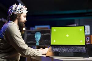 Focus on green screen laptop used by programmer using EEG headset to upload mind into PC. Close up of mockup notebook on desk next to man experimenting with transferring consciousness into cyberspace photo