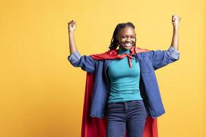 African american superhero flying with red cape, isolated over studio background, flexing muscles. Woman wearing cloak posing as hero in costume showing courage and strength photo