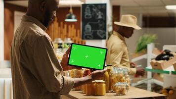 African american buyer shows tablet with greenscreen template in local grocery store, looking at blank copyspace next to chemicals free items. Regular client holds device with chromakey. Camera 1. photo