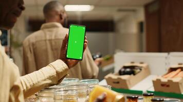 African american storekeeper holding phone with greenscreen in local grocery store, having organic products on sale. Vendor presents isolated chromakey display on smartphone, mockup layout. Camera 1. photo