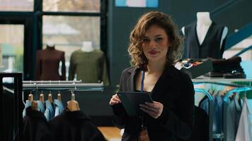 Woman employee examining racks of clothes for stock inventory, using tablet to check all products. Retail store assistant checking discount list on device, ensuring commercial tags. Camera A. photo