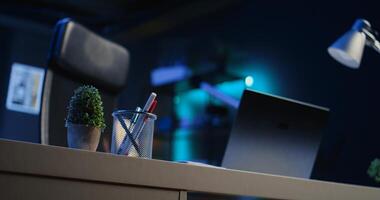 Office desk filled with productivity items in empty home interior illuminated with neon lights at night. Laptop on table and other work related objects in apartment room, camera B close up shot photo