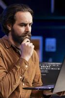 Pensive cybersecurity expert looking confused while using laptop to fix company security vulnerabilities. Contemplative developer feeling worried about potential cyberattacks, close up shot photo