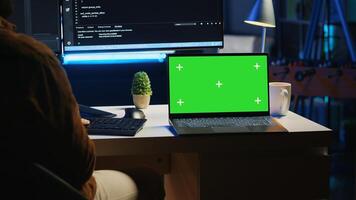 Green screen laptop next to freelancing man doing IT support job from apartment office, coding on PC. Mockup notebook on desk where software engineer writes lines of code on computer at home, camera A photo
