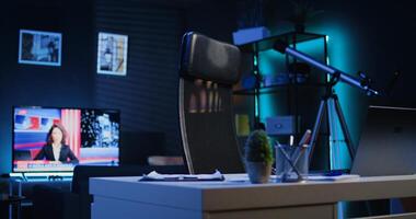 Dimly lit apartment with office desk in front of smart TV displaying news program broadcasting. Laptop on table and television screen in empty room showing channel, camera B close up shot photo