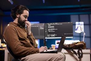 IT specialist working from home using laptop, testing and deploying programs and systems. Programmer checking code displayed on computer monitors, sitting at desk chair in apartment photo