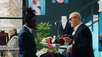 Experienced famous stylist buying the perfect tie color for his rich client, looks for expensive materials at shopping center. Senior fashion designer giving orders to store woman employee. Camera A. photo
