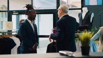Senior designer teaching his apprentice to match ties on elegant suits, presenting the color mixing theory to his pupil. African american man trying to learn basics in fashion industry. Camera A. photo