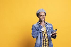 Mature relaxed person thinking about an answer to text on smartphone, looking at his messages on social media app against yellow background. Confident masculine model using mobile phone. photo
