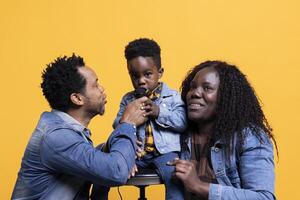 Cheerful adorable small child singing a song on microphone in studio, parents smiling and dancing on his music. Proud mother and father having fun with their happy son on camera. photo