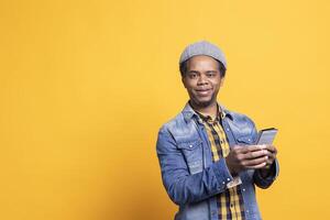 African american male model posing and browsing internet on his phone, standing against yellow background in cool trendy clothes. Happy confident man uses social media apps on camera. photo