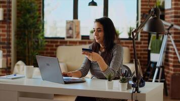 Smiling woman stoked on doing online shopping, carefully typing debit card information on laptop keyboard. Person adding payment method on website while in modern apartment living room, camera B photo