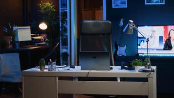 Office desk filled with business productivity items in empty home interior with TV left open in background at night. Laptop, headphones, plants and work documents on table in apartment room, camera A photo