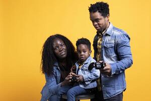 Young african american dad playing with his kid on joystick, using games competition online to have fun together in studio. Smiling parents enjoying time with their toddler. photo