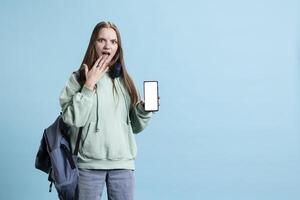 Woman having shocked look on face while holding mockup mobile phone, feeling intrigued by technology. Young girl gasping while presenting isolated screen cellphone, studio backdrop photo