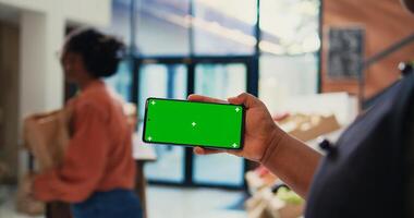 Local merchant showing greenscreen layout on phone, holding device with isolated copyspace template at cash register. African american retailer with smartphone running chromakey display. Tripod shot. photo