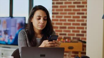 Smiling person checking mobile phone notifications at computer desk, taking break from work. Indian woman in cozy apartment pausing job tasks to read messages on cellphone, camera B close up photo