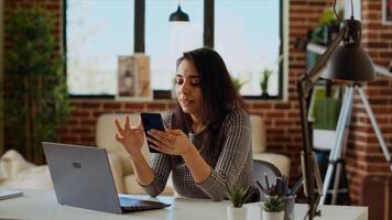 Teleworker checking her phone notifications at home in front of laptop, taking break from work. Woman in cozy apartment office taking pause from job tasks to read messages on smartphone, camera B photo