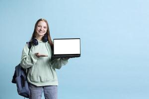 Upbeat girl pointing towards laptop with isolated screen display, doing advertisement, studio background. Radiant teenager grinning, creating ad promotion with mockup notebook device photo