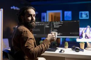 Portrait of happy computer technician enjoying hot tea while implementing security measures to protect company systems and data. Joyful IT worker holding coffee mug and debugging code photo