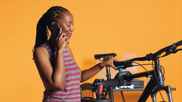 Woman talking on phone with technician, asking for help on fixing defective bike, studio background. BIPOC person on smartphone call with repairman, needing to recondition broken bicycle, camera A photo