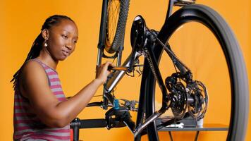 BIPOC engineer checking bicycle performance by spinning pedals. Skilled specialist ensuring proper function of tires on bike wheels in orange studio background during maintenance, camera B photo