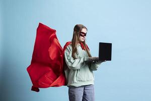Portrait of happy teenager with mask typing on laptop, portraying confident superhero, studio backdrop. Jolly heroic person posing as powerful hero character in costume sending mails on notebook photo