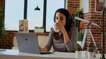 Teleworker checking emails on laptop while at computer desk, drinking coffee and listening music. Remote employee with headphones on working from home solving tasks, enjoying hot beverage, camera B photo