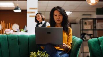 Asian tourists looking for landmarks to visit during their stay, using laptop and tablet while they wait for their room at hotel. Two girlfriends relaxing in lounge area with devices. Handheld shot. photo