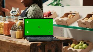 Laptop with greenscreen display running in local grocery store, on counter filled with bulk products stored in reusable glass jars. Isolated mockup chromakey layout on wireless pc at farmers market. photo